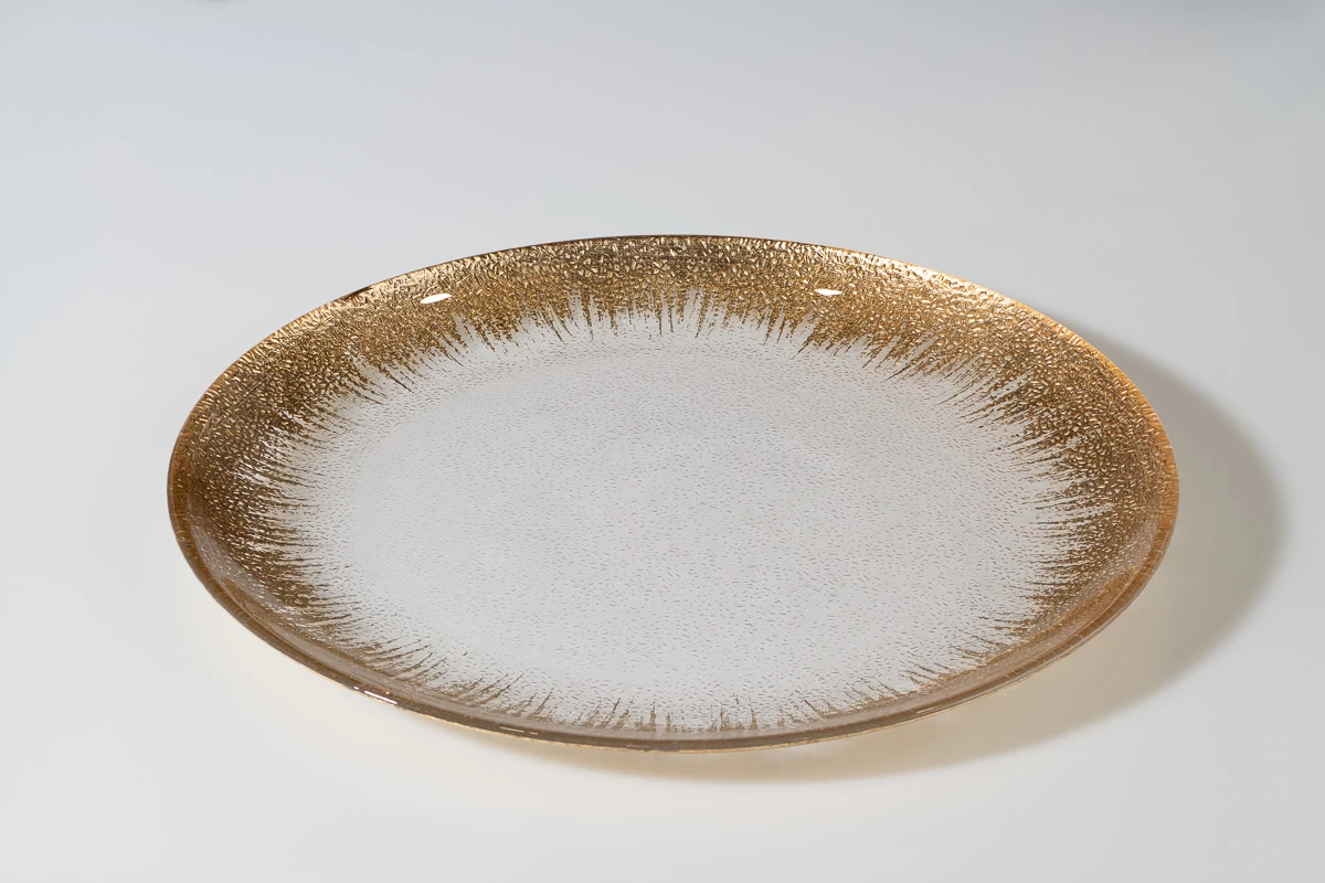 צלם סטודיו מציג צלחת זכוכית דקורטיבית עגולה עם עיטור זהב ייחודי. צילום מוצר לאינטרנט, רקע לבן.